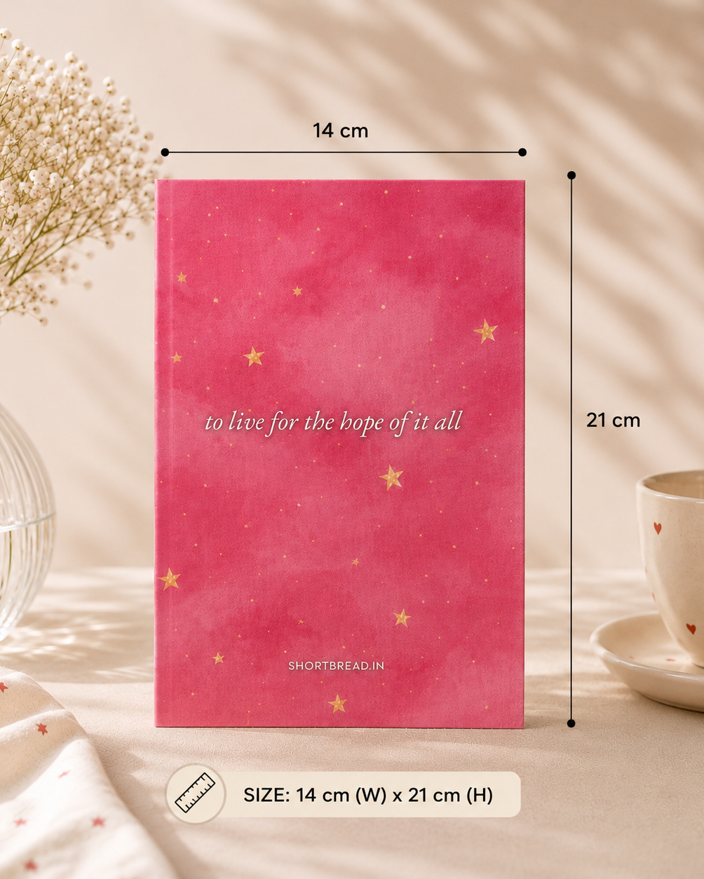 A pink notebook with gold stars and the text 'to live for the hope of it all' by SHORTSBREAD.IN, shown with dimension markers indicating 14 cm width and 21 cm height, on a styled beige background with a vase of flowers and a cup.