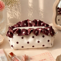 A satin cosmetic pouch with a ruffled burgundy top trim and white polka dots, styled on a tabletop with flowers, a mirror, pearls, and beauty accessories.