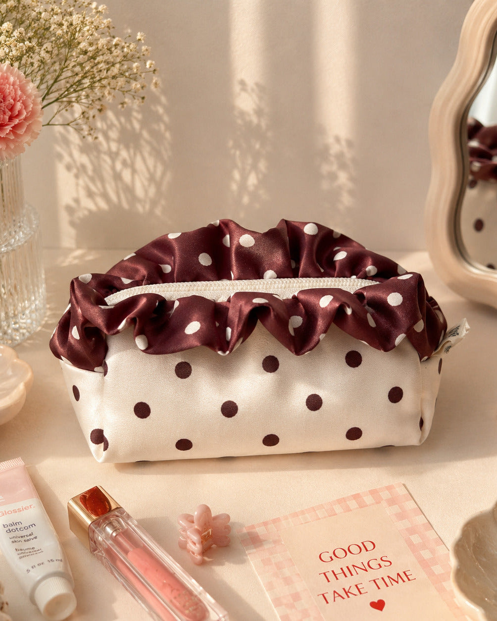 A satin cosmetic pouch with a ruffled burgundy top trim and white polka dots, styled on a tabletop with flowers, a mirror, pearls, and beauty accessories.